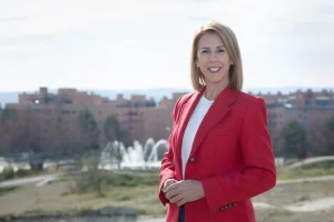 Una mujer con el pelo rubio liso hasta los hombros, que lleva una chaqueta roja y un top blanco, posa al aire libre frente a un paisaje urbano, captada en un estilo «borrador automático», con edificios, árboles y una fuente al fondo.