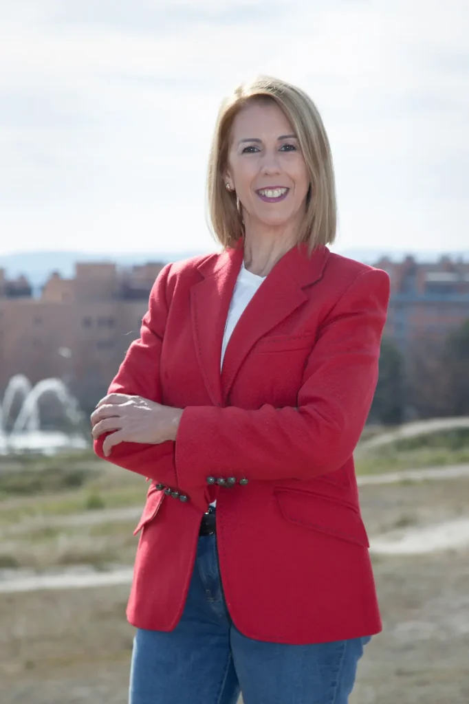 Una mujer con el pelo rubio liso hasta los hombros está de pie al aire libre con los brazos cruzados; lleva una chaqueta roja, un top blanco y unos vaqueros azules. En el fondo, difuminado, se ven edificios y una fuente en PP Rivas, donde Janette Novo hace campaña para 2027.