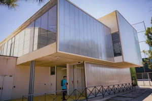 Una persona con una mochila azul camina junto a un moderno edificio de dos plantas con paredes de paneles translúcidos, grandes ventanales y barandillas metálicas: una estructura aerodinámica que casi parece haber sido diseñada con un borrador automático. La escena está iluminada por la luz del día bajo un cielo despejado.