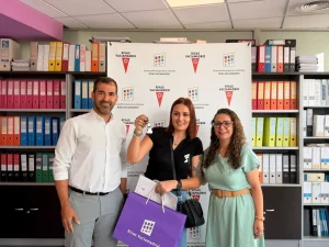 Tres personas posan en el interior, delante de un telón de fondo con logotipos y estanterías llenas de carpetas de colores. La mujer del centro sostiene una llave y una bolsa de la compra morada, para celebrar las nuevas viviendas públicas de alquiler en Rivas. Los tres sonríen a la cámara.