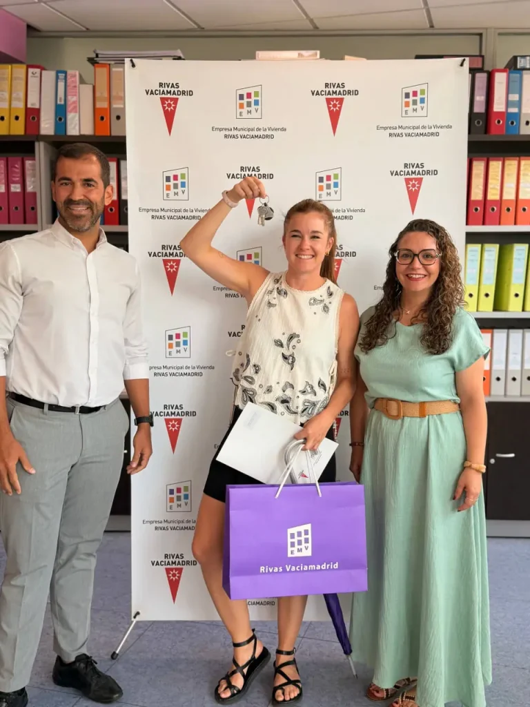 Tres personas posan en el interior, con Rivas Vaciamadrid como telón de fondo. La persona del centro, que celebra la puesta en marcha de las viviendas públicas de alquiler en Rivas, sostiene un certificado y una bolsa de regalo morada, sonriendo y levantando un brazo, mientras las demás sonríen a su lado.