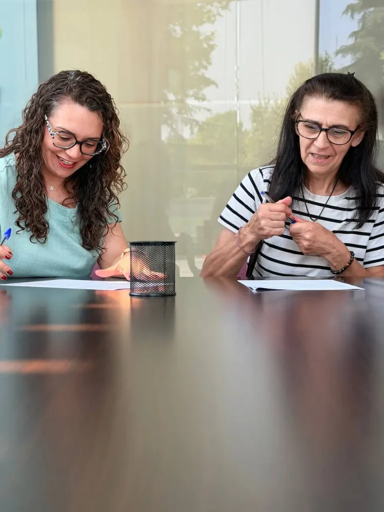 Dos mujeres están sentadas en una mesa larga en Rivas, cada una escribiendo en un papel. Una lleva una camiseta azul claro y gafas, y la otra, una camiseta a rayas y gafas. Entre ellas hay un portalápices de malla negra mientras hablan sobre viviendas públicas y opciones de alquiler.