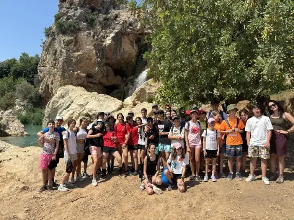 Un grupo de niños, jóvenes y adultos posa para una foto al aire libre, cerca de unas grandes rocas y una cascada en Granada, rodeados de árboles y vegetación. El grupo parece estar realizando una excursión de senderismo o una salida a la naturaleza.