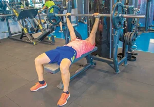 Una persona con camiseta rosa y pantalones cortos azules está tumbada en un banco realizando un press de banca con barra en un gimnasio, lo que refleja el compromiso de los gimnasios de Rivas con la protección de los usuarios. Al fondo se ven diversos aparatos de gimnasio y pesas.