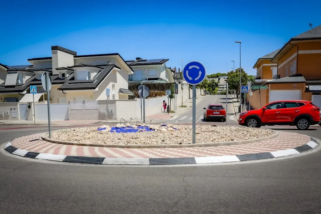 Una pequeña rotonda en la avenida Pilar Miró, con grava en el centro rodeada de ladrillos y baldosas, está rodeada de viviendas; se ven dos coches y un peatón, junto con señales de rotonda y de paso de peatones, lo que refleja un proyecto vecinal bien pensado.