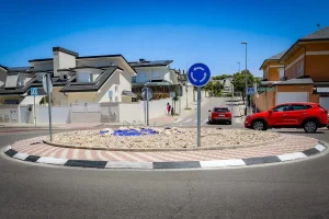 Una pequeña rotonda en la avenida Pilar Miró, con grava en el centro rodeada de ladrillos y baldosas, está rodeada de viviendas; se ven dos coches y un peatón, junto con señales de rotonda y de paso de peatones, lo que refleja un proyecto vecinal bien pensado.
