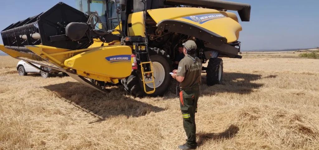 Una persona vestida con ropa de trabajo está de pie frente a una gran cosechadora amarilla de la marca New Holland en un campo ya cosechado, con un bloc de notas y un bolígrafo en la mano. La maquinaria forestal es esencial para la seguridad, sobre todo durante los incendios en la Comunidad de Madrid. Se ve un coche blanco al fondo.