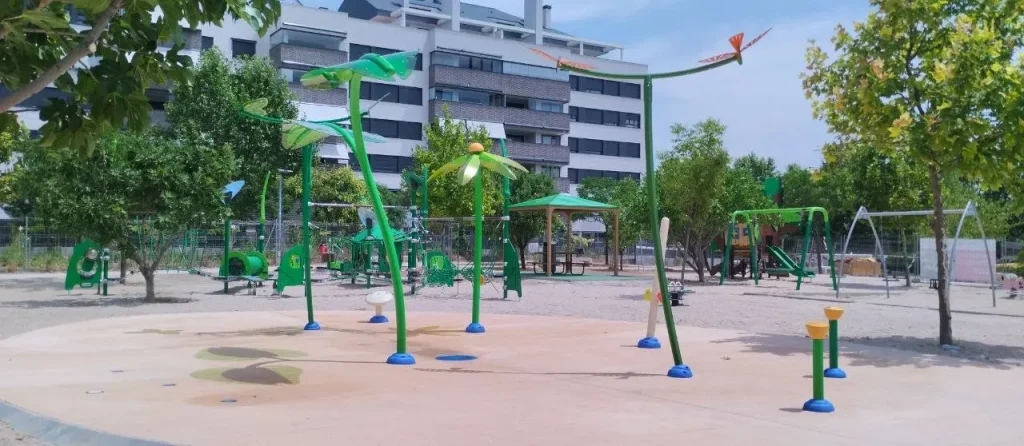 Parque infantil al aire libre con estructuras de juego de colores verde y azul, con juegos de agua, situado en una zona de arena y rodeado de árboles, con edificios residenciales al fondo. En el Parque del 8M también se pueden ver columpios y estructuras para trepar.