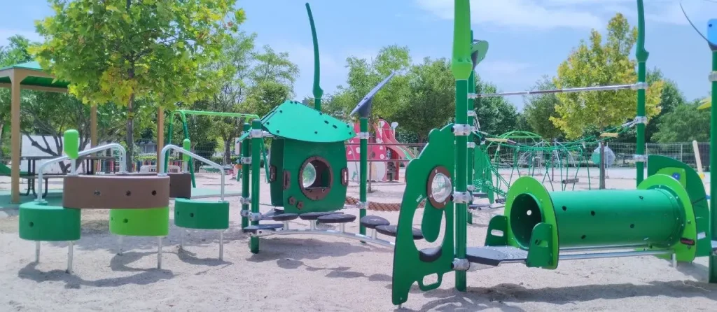 Un moderno parque infantil al aire libre, con estructuras verdes para trepar y toboganes, se encuentra sobre una superficie de arena, rodeado de árboles y vallas, creando nuevos espacios infantiles donde los niños pueden disfrutar de su infancia. Al fondo se divisan otros elementos de juego de colores vivos, que reflejan el espíritu vibrante de Rivas.