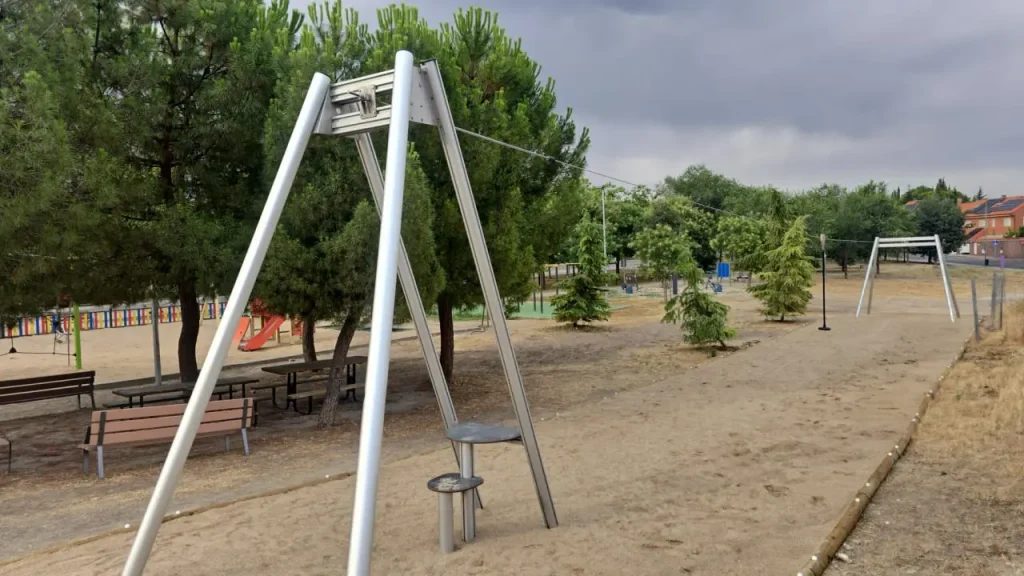 En una zona arenosa de un parque de Rivas se alza una tirolina con un asiento circular, que ofrece nuevos espacios para que los niños jueguen y exploren, rodeada de bancos, árboles y un cielo nublado sobre sus cabezas. Al fondo se divisa otra estructura similar.