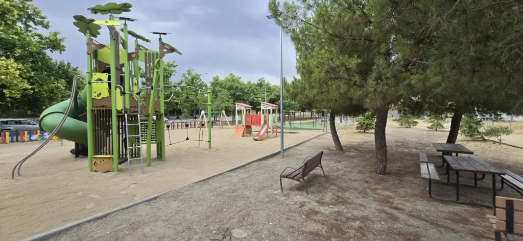 Un parque infantil con toboganes, estructuras para trepar y columpios se encuentra sobre arena: uno de los «espacios para la infancia» cuidadosamente diseñados en Rivas. Cerca hay un banco y una mesa de picnic, a la sombra de los árboles. La zona está vallada y rodeada de vegetación, perfecta para que los niños exploren estos «nuevos espacios». El cielo está nublado.