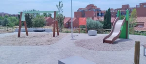 Un parque infantil con un columpio y un tobogán sobre suelo de grava ofrece espacios para los niños en Rivas. Al fondo se ve un cartel naranja, mientras que detrás de una valla de madera se divisan edificios residenciales y árboles. El cielo está parcialmente nublado.