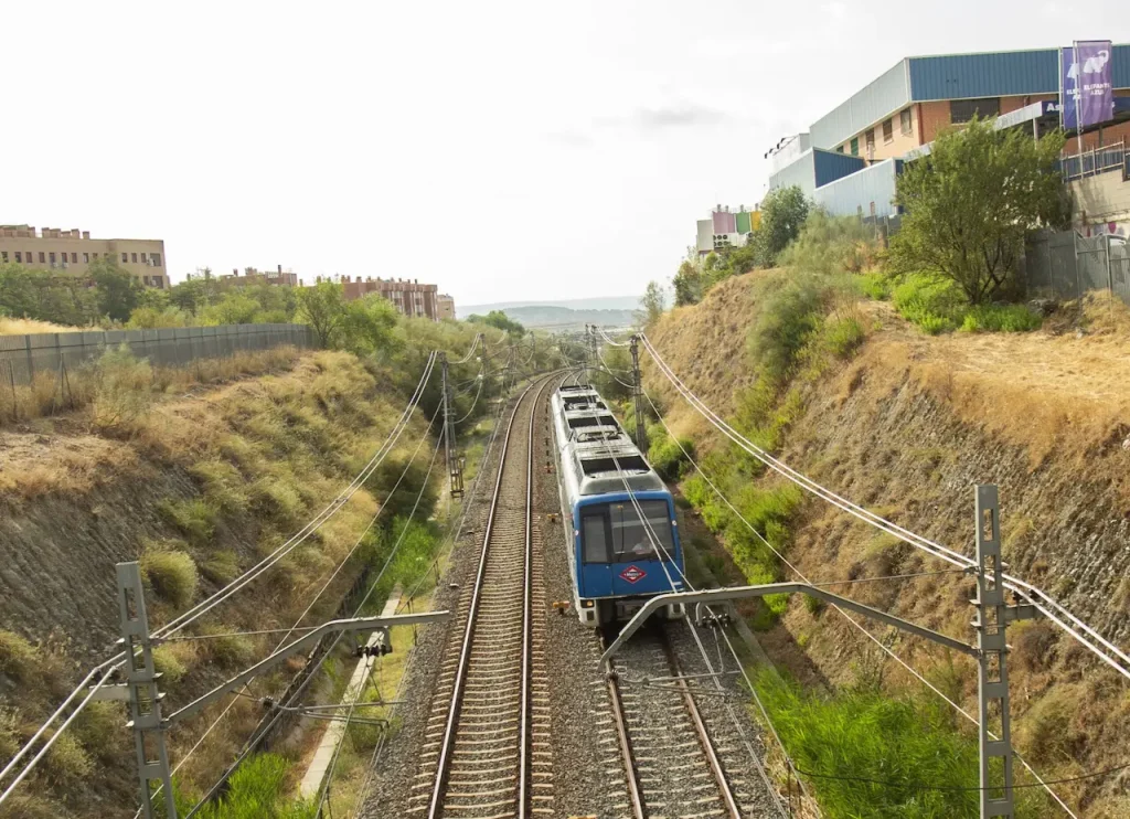 Un tren de cercanías azul circula por una vía doble a través de un desmonte seco y cubierto de hierba entre colinas, pasando junto a viviendas y edificios a ambos lados bajo un cielo despejado, con cables eléctricos que se extienden por encima.