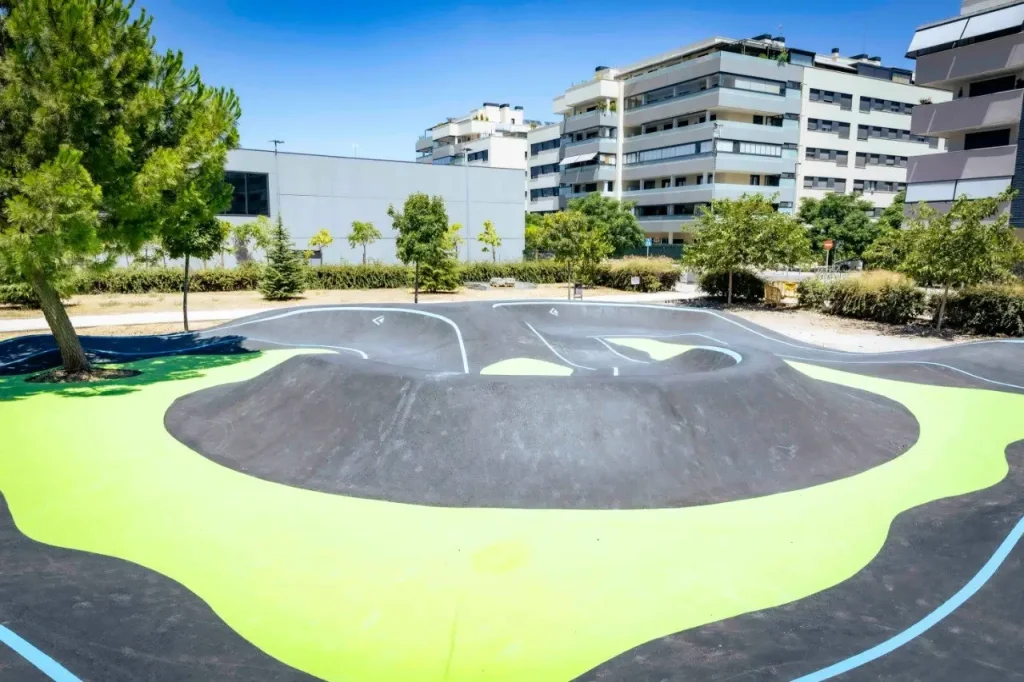 Una pista de pump track al aire libre, con marcas negras y verdes, está rodeada de árboles y modernos edificios de apartamentos, con un cielo azul y despejado al fondo, creando nuevos espacios en Rivas perfectos para los niños.