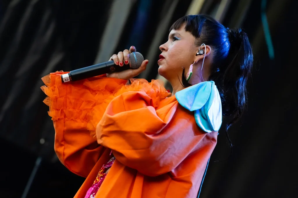 Una mujer vestida con un traje naranja de mangas anchas con volantes canta ante un micrófono en el escenario, con un auricular en la oreja y un fondo oscuro a sus espaldas.