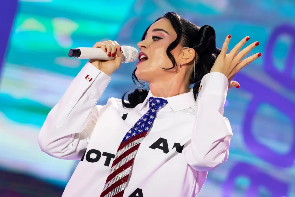 Una persona canta ante un micrófono en el escenario; lleva una camisa blanca con letras negras y una corbata con el diseño de la bandera estadounidense. Tiene las uñas pintadas de rojo y el pelo oscuro recogido en dos coletas altas. El fondo es colorido y está desenfocado.