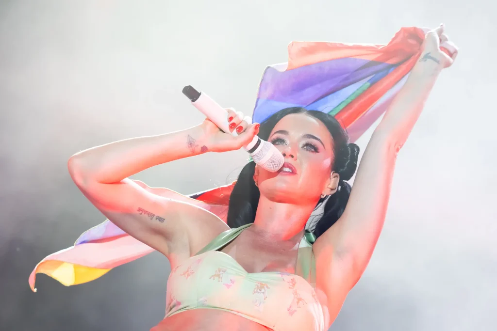 Una mujer que canta ante un micrófono blanco sostiene una bandera arcoíris por encima de la cabeza. Lleva una camiseta de color verde claro y está de pie frente a un fondo lleno de humo y muy iluminado.