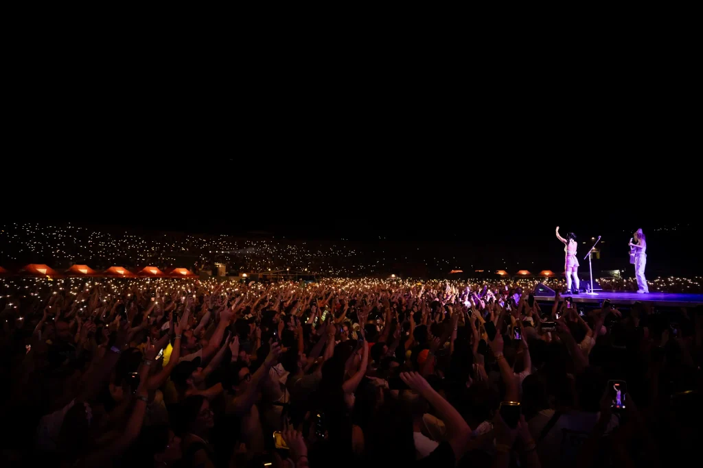 Una gran multitud en un concierto al aire libre levanta las manos y los móviles hacia un escenario intensamente iluminado, donde un artista se encuentra bajo los focos en plena noche. El fondo es oscuro, con luces dispersas visibles en la distancia.