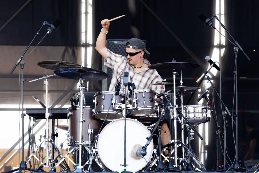 Un baterista que lleva gafas de sol y una gorra toca la batería en el escenario, sosteniendo una baqueta por encima de la cabeza. Hay varios micrófonos colocados alrededor de la batería, y al fondo se ven equipos de escenario e iluminación.