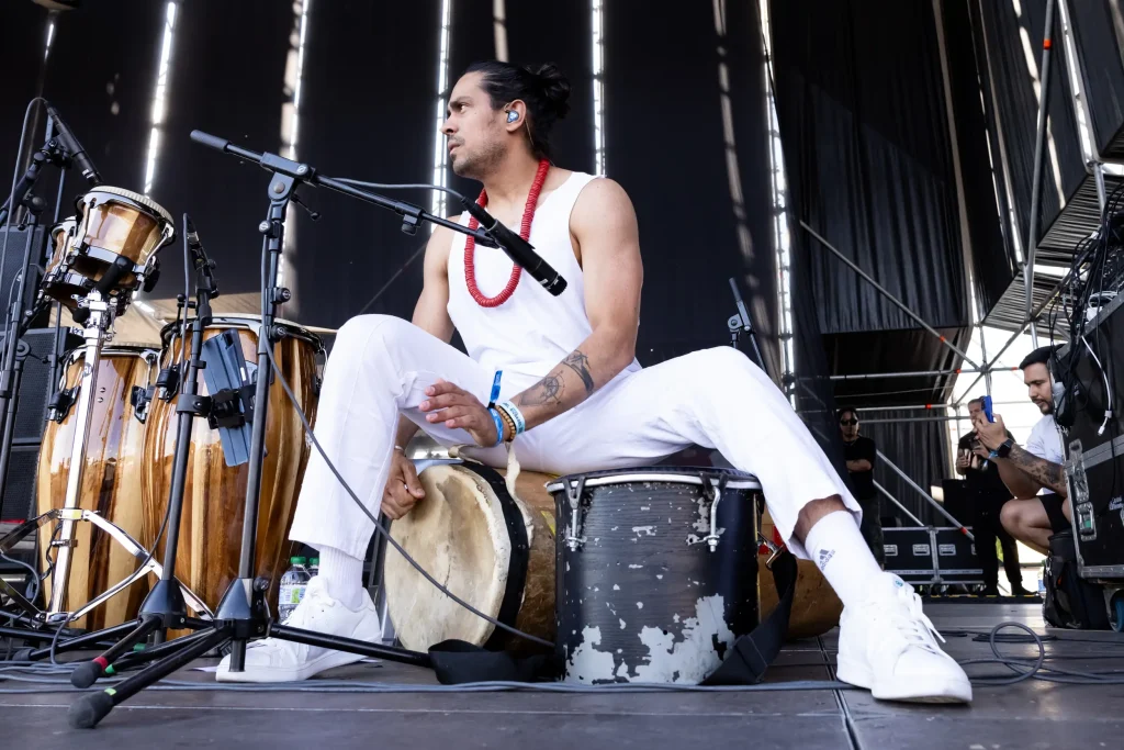Un músico vestido de blanco y con collares rojos está sentado en el escenario, rodeado de tambores, y toca uno de ellos con la mano. Se ven micrófonos y el equipo del escenario, y hay otra persona al fondo.