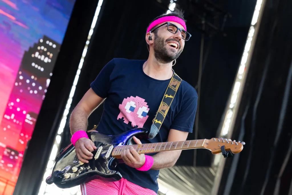 Un hombre con gafas, una diadema rosa y una camiseta negra con un fantasma rosa de Pac-Man toca la guitarra eléctrica en el escenario. Está sonriendo y lleva pulseras y pantalones cortos de color rosa.