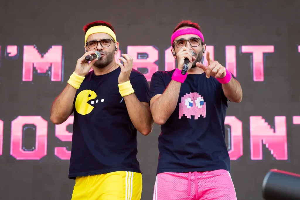 Dos hombres actúan en el escenario con micrófonos. Uno lleva una camiseta de Pac-Man y unos pantalones cortos amarillos; el otro, una camiseta con un fantasma de Pac-Man y unos pantalones cortos rosas. Ambos llevan cintas para el sudor. En el fondo, una pantalla digital muestra un texto.