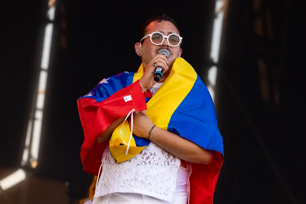 Una persona vestida de blanco y con unas gafas blancas grandes está cantando en el escenario, envuelta en una bandera venezolana y con un micrófono en una mano.