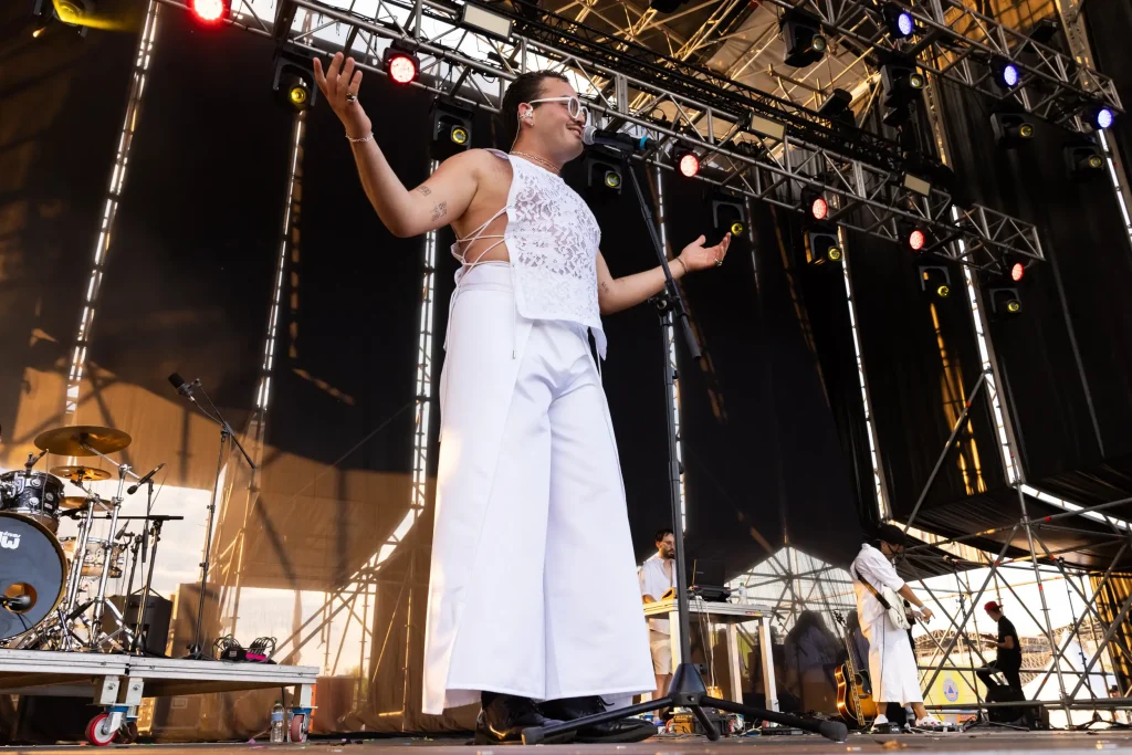 Una persona que lleva un top blanco de encaje sin mangas y unos pantalones blancos de corte ancho está de pie en un escenario al aire libre con los brazos abiertos. Al fondo se ven músicos e instrumentos, y las luces del escenario brillan sobre su cabeza.