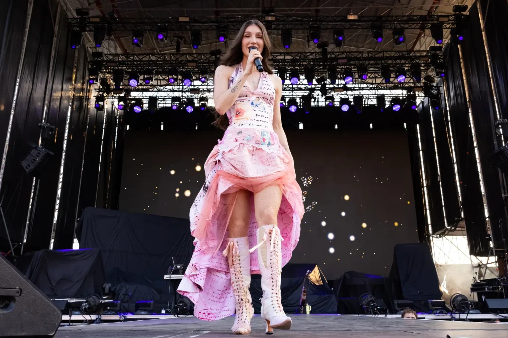Una mujer con un vestido rosa de capas y botas con cordones está de pie en el escenario de un concierto al aire libre, sosteniendo un micrófono. Detrás de ella se ven las brillantes luces del escenario y una gran pantalla con efectos de luz.