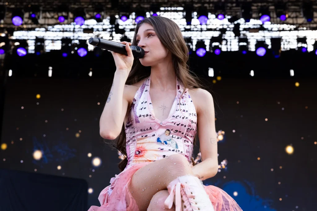 Una mujer con el pelo largo y castaño canta ante un micrófono en un escenario al aire libre; lleva un vestido sin tirantes de color claro con letras e imágenes, y unas botas blancas. Al fondo se ven luces de escenario azules y amarillas.
