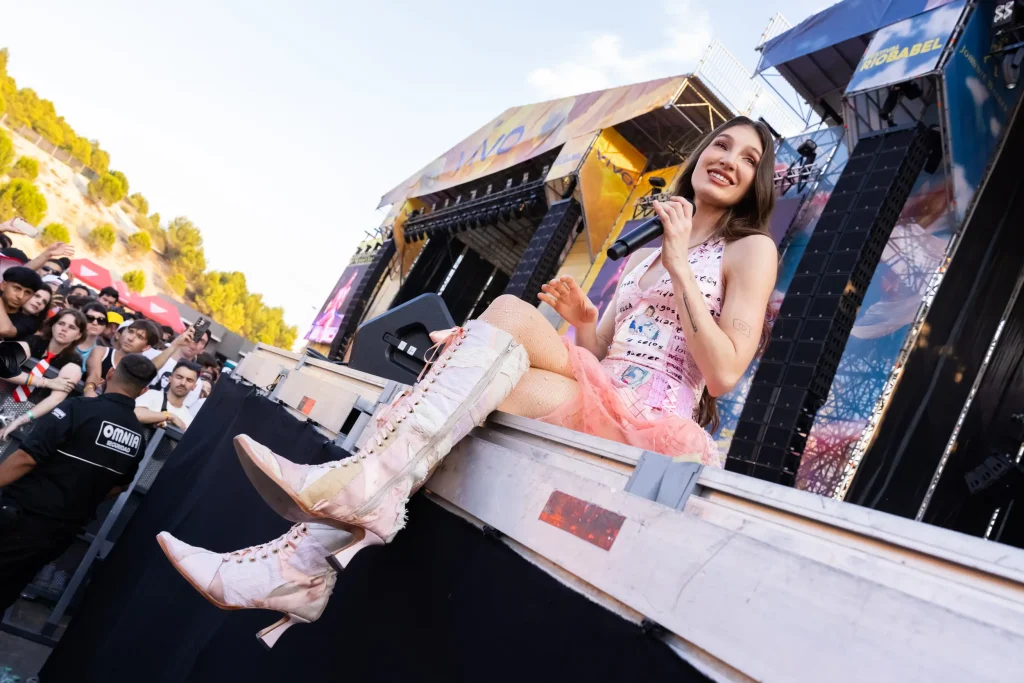 Una artista con un vestido rosa y botas altas está sentada en el borde del escenario de un concierto, sonriendo y con un micrófono en la mano, con el público y el personal de seguridad visibles al fondo.