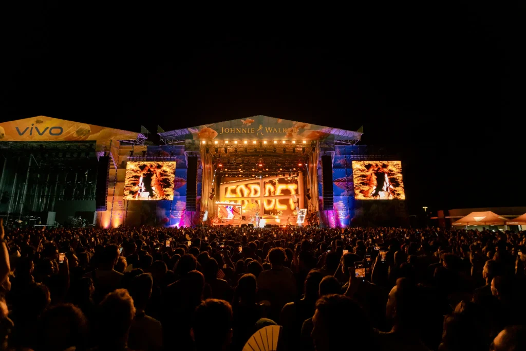 Una gran multitud contempla por la noche un escenario de concierto al aire libre muy bien iluminado, con dos pantallas gigantes a ambos lados en las que se proyectan imágenes y, en las pancartas del escenario, se pueden ver las palabras «Johnnie Walker» y «Vivo».