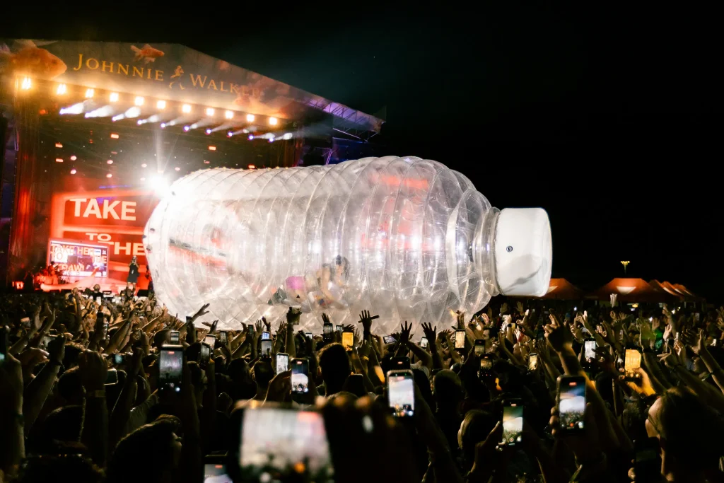 Una gran multitud asistente a un concierto nocturno sostiene una botella hinchable gigante con gente dentro, mientras que al fondo se ve un escenario intensamente iluminado con el letrero de JOHNNIE WALKER.
