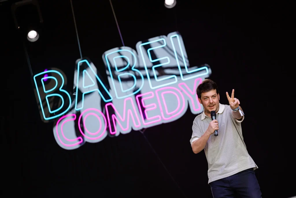 Un hombre que sostiene un micrófono está de pie en el escenario, delante de un gran letrero de neón en el que se lee «Babel Comedy». Está haciendo el signo de la paz con la mano.