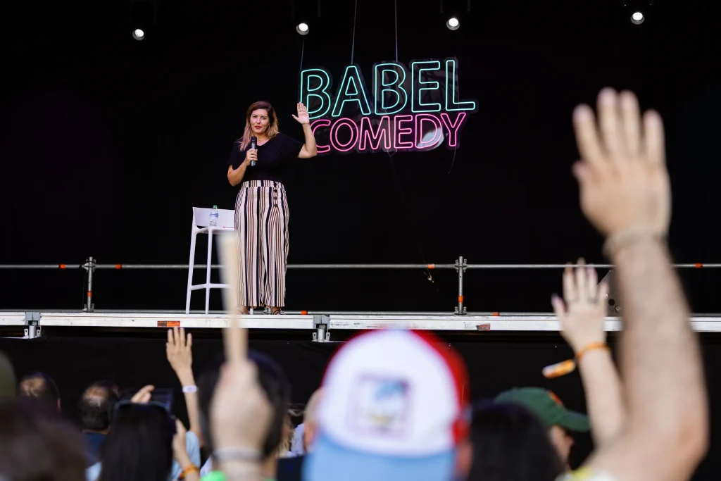 Una mujer está de pie en el escenario, sosteniendo un micrófono frente a un letrero de neón que dice «Babel Comedy», con miembros del público levantando la mano en primer plano. Junto a ella hay un taburete blanco con una botella de agua.