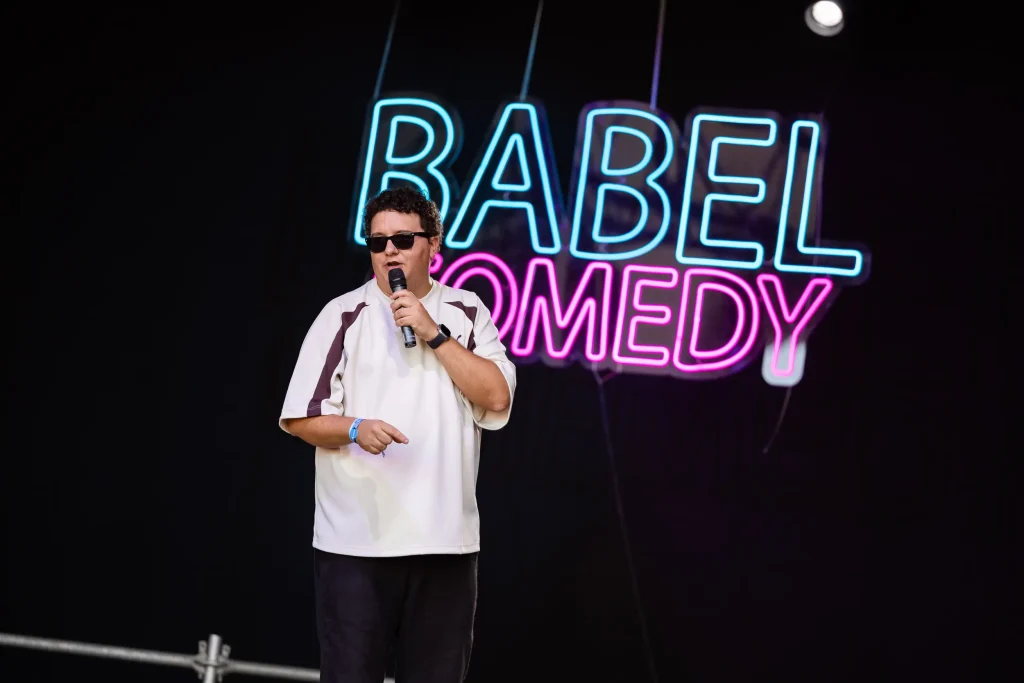 Un hombre con gafas de sol y camisa blanca habla por un micrófono en el escenario. Detrás de él, un letrero de neón dice «Babel Comedy» en letras azules y rosas sobre un fondo negro.