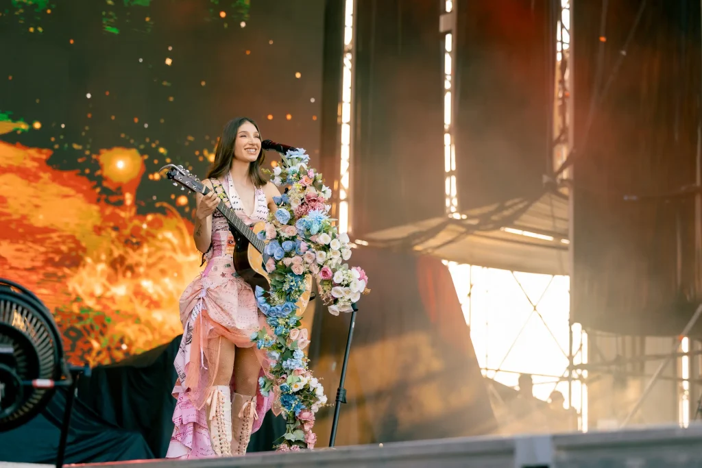 Una música vestida con un vestido rosa actúa en el escenario con una guitarra decorada con flores de colores. Al fondo se ven las brillantes luces del escenario y una gran pantalla con imágenes abstractas.