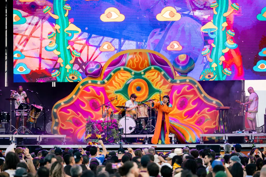 Una banda actúa en el escenario frente a un colorido telón de fondo psicodélico con grandes motivos de ojos. El cantante lleva un traje naranja. El público asiste al concierto y se ven varios instrumentos en el escenario.