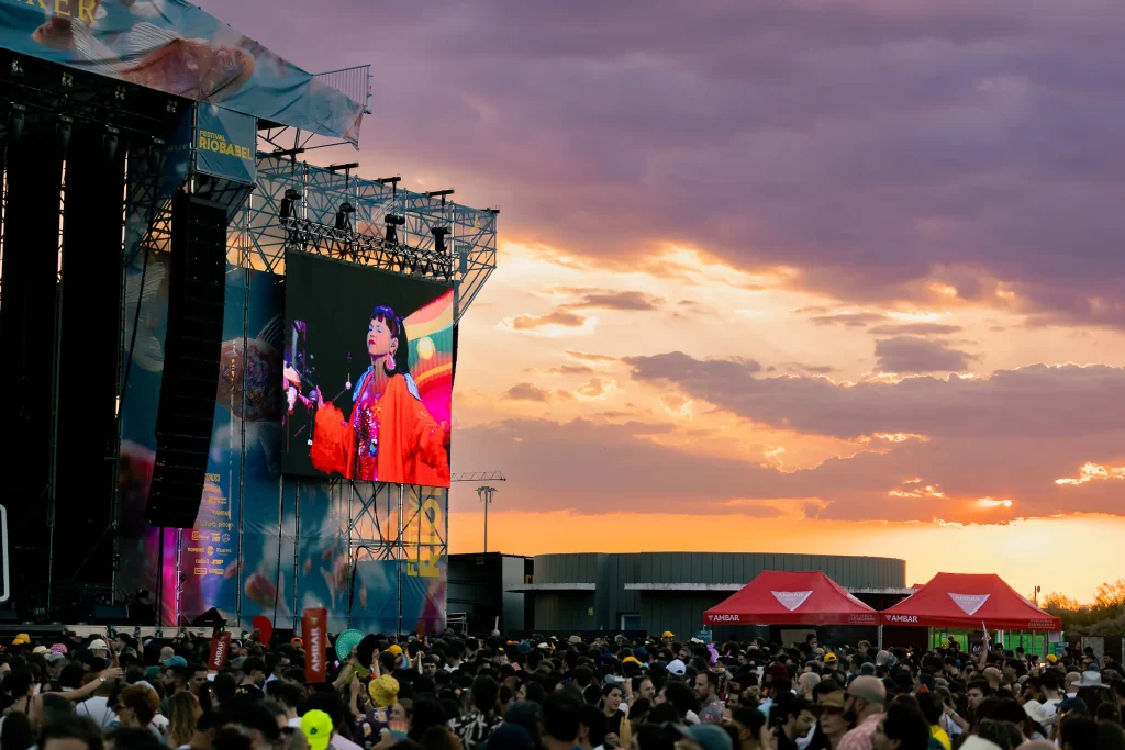 Una gran multitud se reúne en un concierto al aire libre al atardecer, mientras se ve a un artista en una gran pantalla situada junto al escenario. Al fondo se divisan dos carpas rojas bajo un cielo lleno de colores.
