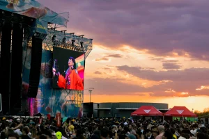 Una gran multitud se reúne en un concierto al aire libre al atardecer, mientras se ve a un artista en una gran pantalla situada junto al escenario. Al fondo se divisan dos carpas rojas bajo un cielo lleno de colores.