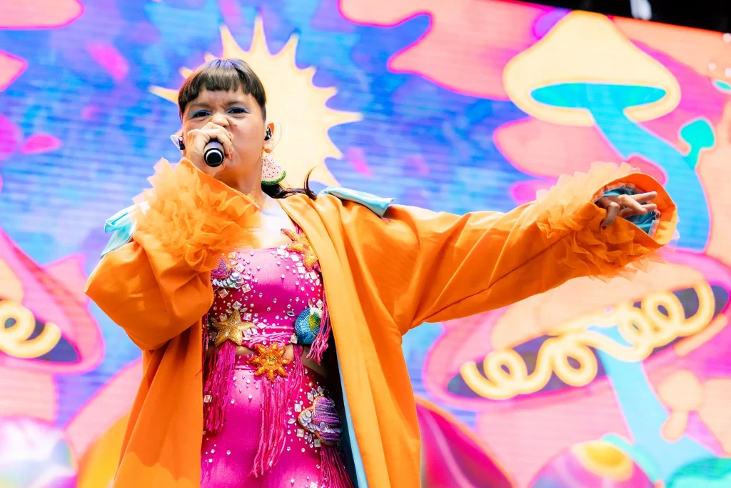Una cantante, vestida con un traje de color rosa brillante y naranja, actúa en el escenario con un micrófono en la mano. El colorido fondo presenta formas abstractas y psicodélicas, así como ilustraciones de setas.