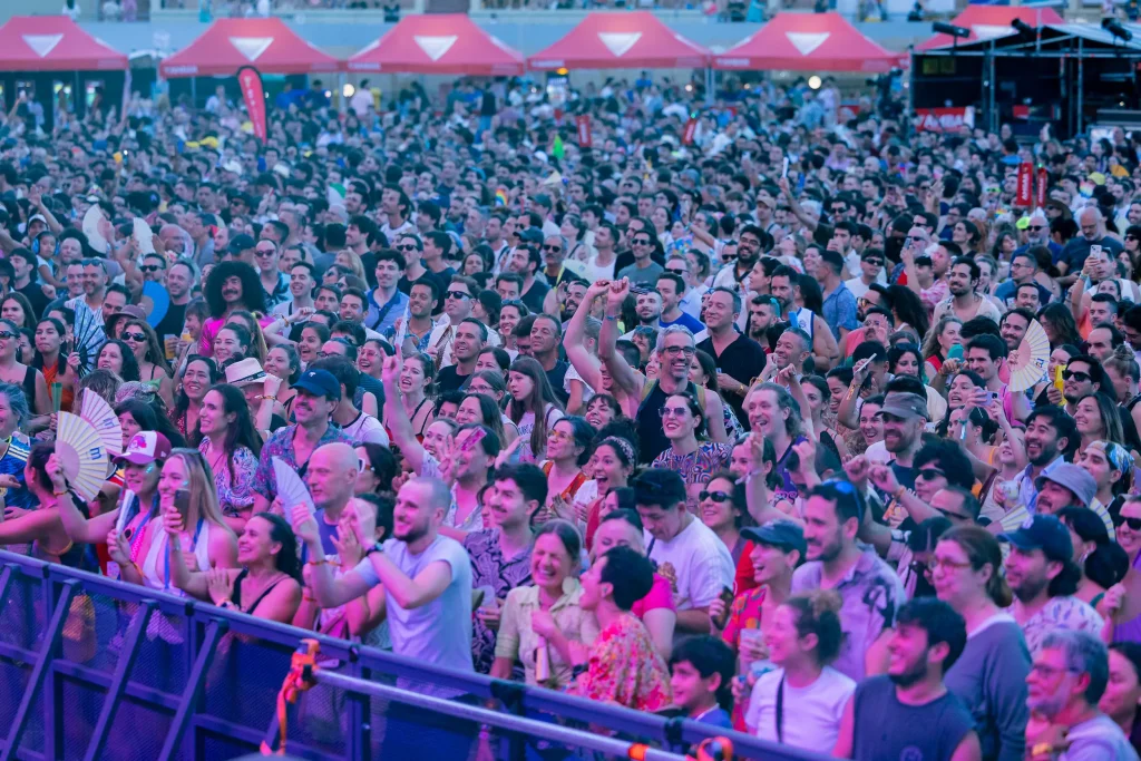 Una gran multitud de personas se apiña en un evento al aire libre o un concierto; muchas de ellas aplauden, vitorean, sonríen y levantan las manos. Al fondo se ven toldos rojos.