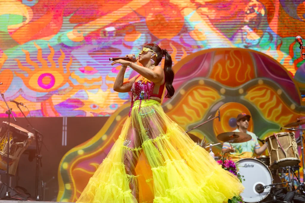 Una cantante, con una falda colorida, transparente y de color amarillo y verde, actúa en el escenario con un micrófono. Detrás de ella, un baterista toca la batería. El fondo presenta motivos psicodélicos muy llamativos y colores vivos.