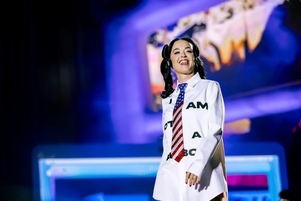 Un artista está de pie en el escenario con una camisa blanca con letras negras y una corbata con motivos de la bandera estadounidense. Al fondo se ven luces brillantes y coloridas y una pantalla borrosa.
