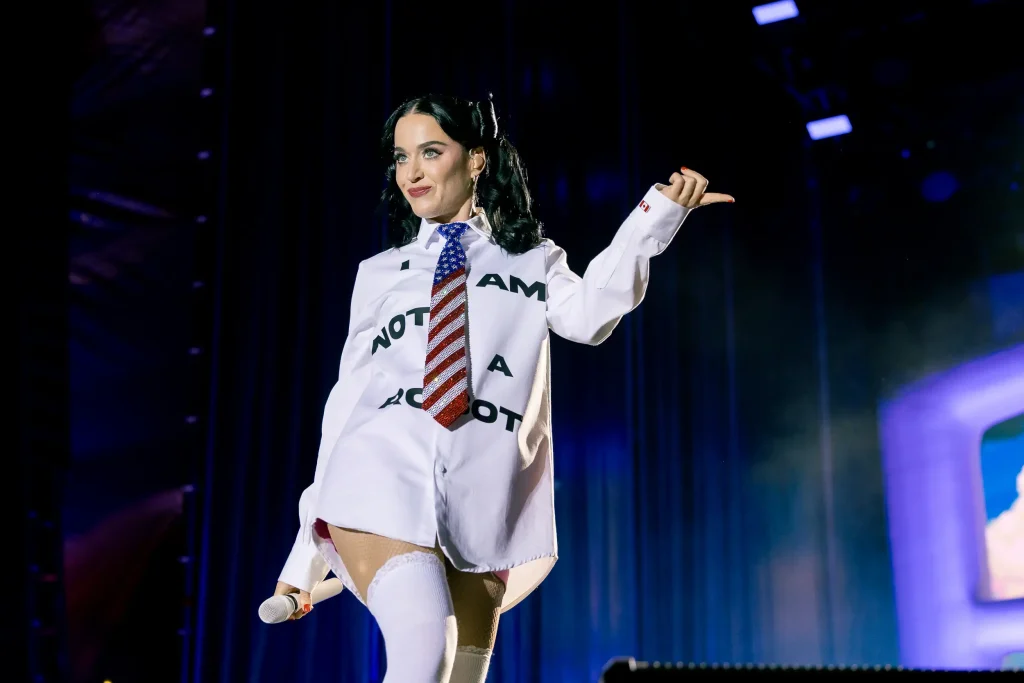Un artista en el escenario lleva una camisa blanca con la inscripción «I AM NOT A ROBOT», una corbata a rayas y medias hasta el muslo, y sostiene un micrófono. Al fondo se ven las luces del escenario y una gran pantalla.
