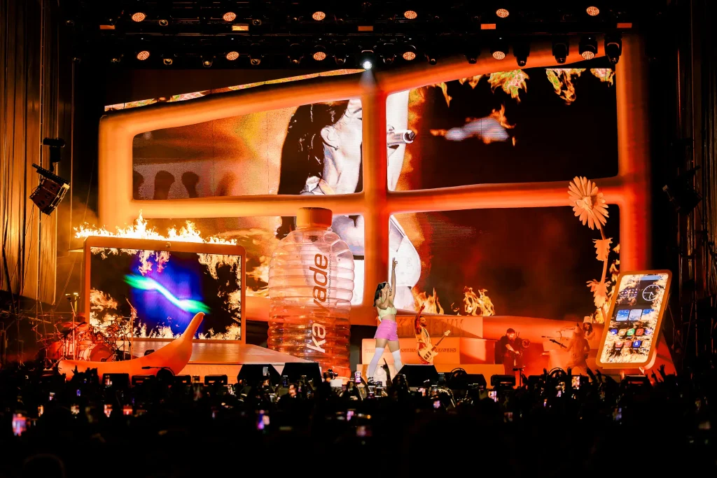 Un cantante actúa en el escenario rodeado de grandes y coloridos elementos escénicos, entre los que se incluyen un móvil gigante, pantallas digitales y atrezo con llamas, mientras el público graba el evento con sus móviles.