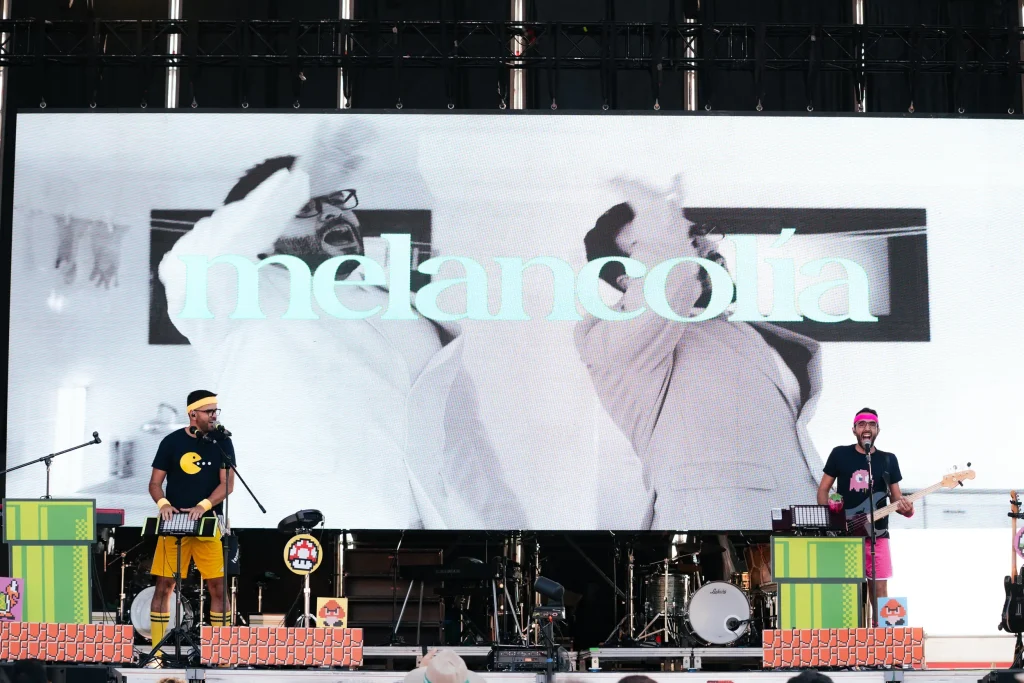 Dos músicos actúan en el escenario frente a una gran pantalla en la que se ve una imagen en blanco y negro de dos personas y la palabra «melancolía» escrita en azul claro. El escenario está decorado con elementos de videojuegos retro.