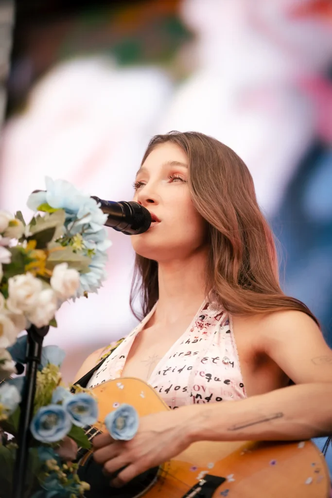 Una mujer con el pelo largo y castaño canta ante un micrófono decorado con flores azules y blancas. Sostiene una guitarra y lleva una camiseta clara con letras, mientras actúa en el escenario.