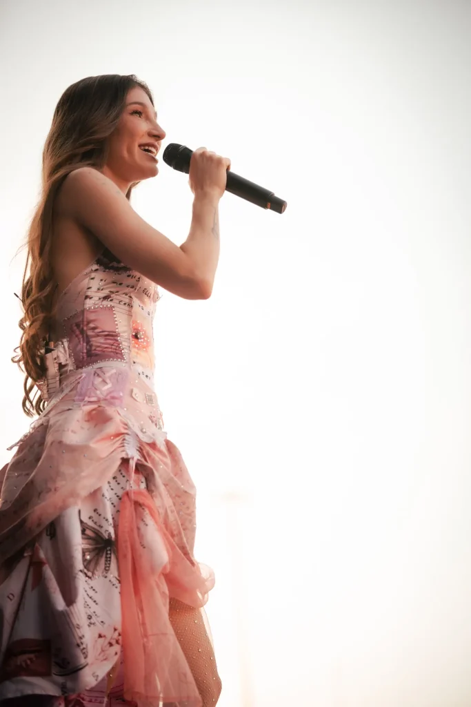 Una persona con el pelo largo y ondulado sostiene un micrófono y sonríe mientras actúa en el escenario; lleva un vestido rosa claro estampado con volantes, frente a un fondo luminoso y suavemente iluminado.