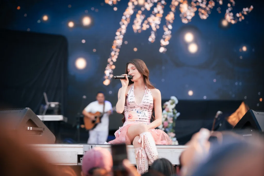Cantante actuando en el escenario con un micrófono, vestida con un vestido sin tirantes de color claro y botas rosas.